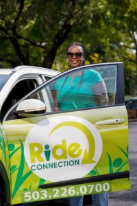 A Ride Connection driver in sunglasses stands by an open car door with the Ride Connection logo.